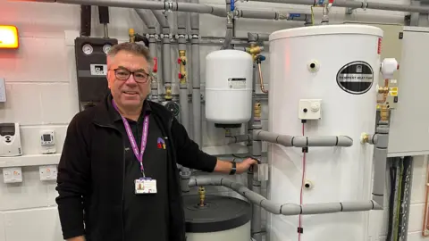 Edd Smith/BBC Colin Hyde standing next to equipment used to help with the training of heat pump installation. He is wearing glasses, along with a cardigan over a black T-shirt.