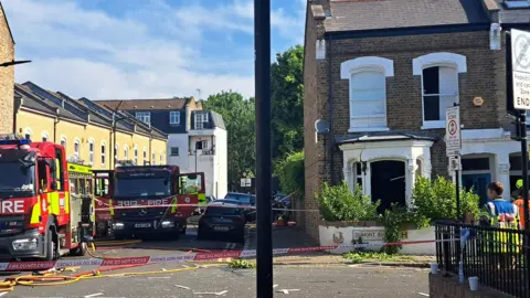PA Media Fire engines and emergency workers outside a damaged brick house on Dumont Road. The front bay window of the house has been blown out, with debris scattered on the street. The area is cordoned off.