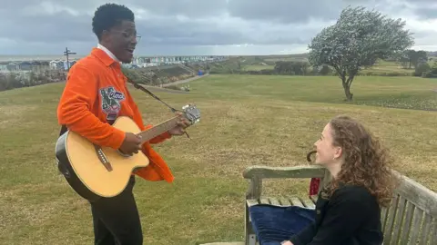 Amy Trigg Jordan Benjamin playing a guitar on the Greensward at Frinton-on-Sea. He is looking at Amy Trigg, who is sat on a bench in front of him and smiling.