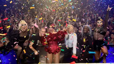 Heaven Nightclub A group of drag queens stand on stage with the owner. Multicoloured confetti can be seen falling over a large crowd in the background. 