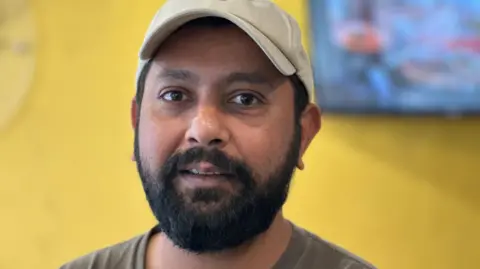 BBC Hassan Ali Tipu, who has a dark beard and it wearing a brown T-shirt and a cream coloured baseball cap.