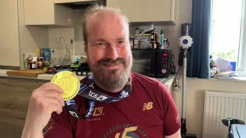 Nathaniel Dye, 39, smiles as he holds a marathon winner's medal around his neck sitting at the kitchen table in his flat