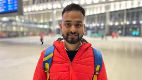 A close up image of Amit Kumar, who is wearing a red jacket and a blue backpack. He is standing in Grand Central Station in Belfast. The background of the image is blurred. 