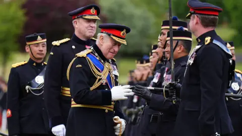 PA Media The King in ceremonial military uniform talks to one of the soldiers as he walks down a line of them. Two people in uniform stand behind him.