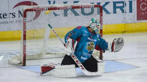 Getty Images A Belfast Giants goaltender in full uniform is in nets. The jersey is teal and the trousers black. The goaltender is holding a large white ice hockey stick in his right hand and trying to catch the puck in a gloved left hand.
