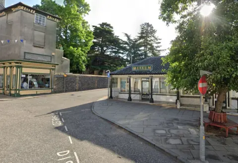 A street level view of Diss Museum, it is shown as being on the corner of a road in Diss with a stone built wall and trees behind it.