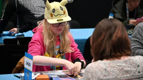 Getty Images Um jovem fã de Pokémon fica do outro lado de uma mesa de um oponente durante uma partida do jogo de cartas comerciais. Usando um boné de beisebol amarelo com orelhas de Pikachu e um capuz rosa brilhante com uma impressão colorida de Pikachu, ela parece atenciosa ao considerar seu próximo passo. 