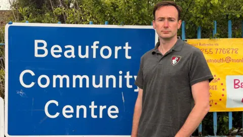 Tom Hayes Bournemouth East MP Tom Hayes, wearing a grey AFC Bournemouth polo, is standing next to a large blue sign for Beaufort Community Centre.