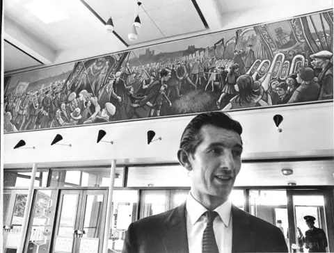 The Story/ Durham County Council Artist Norman Cornish pictured in front of his Durham Miners’ Gala mural in County Hall in 1963. He is clean shaven with thick black hair swept back and he is wearing a suit. The large mural shows people with musical instruments and banners parading.