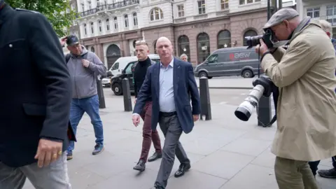 PA Media Ray Connolly with very short light-coloured hair, striding across a pavement wearing a blue jacket, light blue shirt and grey trousers. There is a press photographer with a long lens camera on his arm and a smaller camera up to his eye. Other people are visible and there is a grey van parked on the other side of the road.