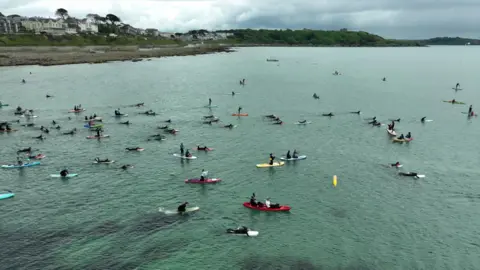 People on paddleboards and canoes in the sea. The water is a light blue. The sky is grey and cloudy. 