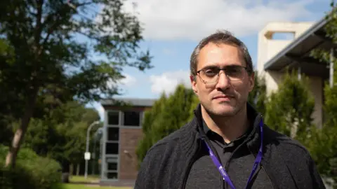 Harper Adams University A man wearing a black shirt and cardigan stands outside on a sunny day, with a purple lanyard around his neck.