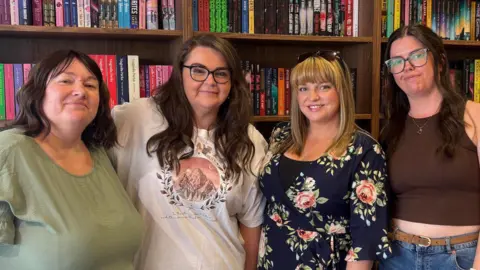 Carrie McGovern, Sara Mack, Ellie White and Hayley Beckinsale. Carrie is wearing a sage green dress. She has shoulder, length brown hair that is curled, with side bangs. Sara has long brown hair that is curled. She is wearing black-rimmed glasses, and a white top that has a design with three mountain peaks within a circle, and the word Velaris above them. Ellie has blonde hair that is medium length with a fringe. She is wearing a floral dress that is navy with pink peonies and roses, and she has a pair of tortoiseshell sunglasses on the top of her head. Hayley has long, dark brown hair, and is wearing tortoiseshell glasses and a brown vest top. They are stood together in front of a bookcase. Each shelf is full with an array of books in different colours and genres. 