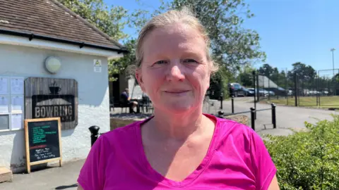 Donna pictured looking into the camera and smiling slightly. She has blonde grey hair tied up and is wearing a hot pink athletic top. She is standing outside the Bowring Park Cafe. It is a single-storey, small building, painted white and a chalkboard sign is standing outside it.
