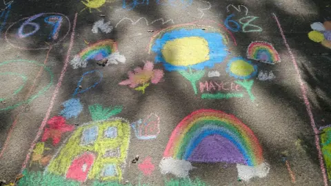 Ross Le Brun Art work on the tarmac drawn in chalk - a pineapple house depicting spongebob squarepants characters bottom left, a rainbow bottom right, another smaller rainbow, flowers and a cloud top left and bigger blue flowers and the name Maycee next to another rainbow top right