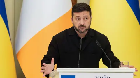 Zelensky looks to the right of frame while gesturing with his hands as he speaks from behind a lecturn with the Ukrainian flag on it, with Ukrainian and Irish flags in the background, in Kyiv on 23 July.