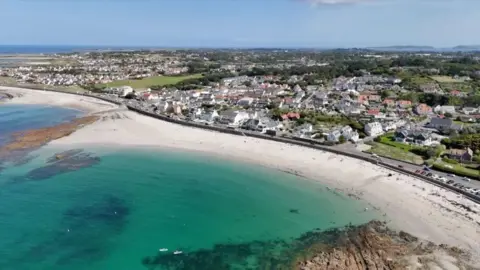 A long beach with rocks, sea and sand with a breakwater and houses and fields behind.