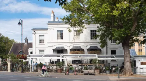 A large white Georgian building is partially obscured by a metal pergola with a retractable roof. 