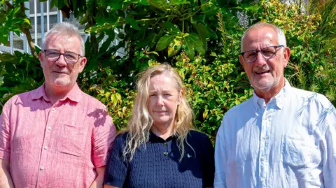 Les Amis A man with glasses in a pink shirt, a woman with blonde hair in a blue top and a man with glasses in a light blue shirt.