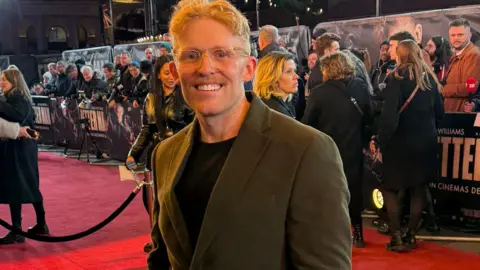 Adam Tucker Adam Tucker stood out at the London red carpet premiere of Better Man wearing a suit blazer and black T-shirt. The fans and press are standing behind them on the other side of the barrier.