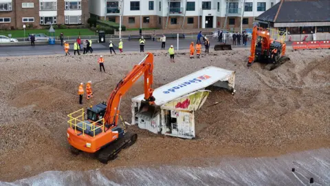 Eddie Mitchell Two orange excavators on the beach removing shingle from a white shipping container so they can move it