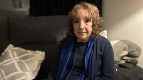 Uma mulher mais velha, com cabelos levemente cacheados, está sentada sorrindo em um sofá cinza. 