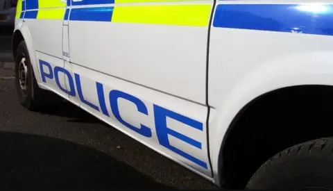 A side view of a police van with a logo spelling out the word police in capital letters above which is a stick-on yellow and blue patterned stripe along with a view of the front wheel and a partial but enticing view of a rear tyre coloured black.