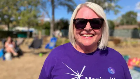 A woman with white hair in a bob wearing black sunglasses and a purple t-shirt is smiling on a summers day.