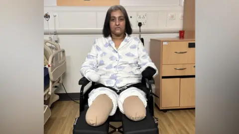 Manjit in a wheelchair wearing patterned pyjamas, with both legs and arms wrapped after they were amputated. The hospital room includes a bed, cabinets and medical fittings.