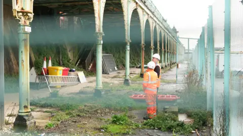 Eddie Mitchell Two men in orange hi-vis uniforms and white hard hats stand at the bottom of the terrace looking up at it