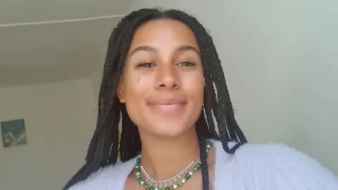 Darwin Arnold Darwin Arnold smiles at the camera while taking a selfie inside a room painted white. She wears a fluffy pale purple cardigan and a green beaded necklace, her braided hair worn loose over her shoulders. 