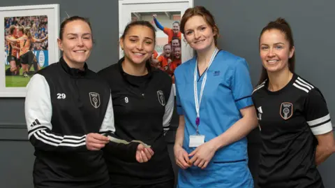NHS Greater Glasgow & Clyde Four women standing facing the camera, while standing indoors. Three of the women are wearing either tracksuits or football tops for the Glasgow City club. The fourth woman is wearing blue nurses scrubs.