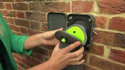 Gray and green phone pouch pressed against a square plastic unlocker mounted on a brick wall