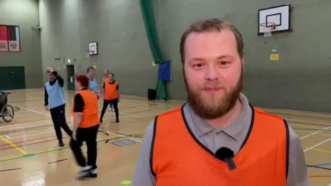 BBC Lee Campbell is wearing a bright orange sports bib inside a sports hall with other people playing sports in the background.
