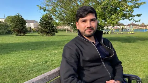 A man is sitting on a bench, smiling. He is wearing a black jacket. 