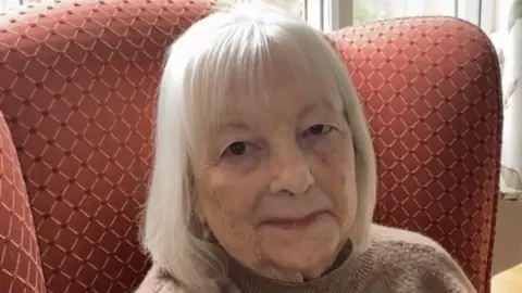 Ellie Bishop Margaret Taylor is smiling and sitting in a red patterned chair. She has a white bob and is wearing a beige jumper.