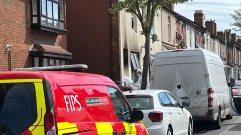 A view of a house with black smoke damage on the walls, cordoned off by police tape. Forensics officers in white suits are entering the house.