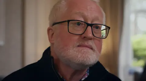 Firecrest Films Headshot of a man with short grey hair, dark-rimmed glasses and a well trimmed beard and moustache. He is wearing a blue and red checked shirt underneath a navy half-zip sweater