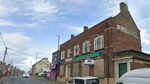 Boarded up pub with green sign saying "The Travellers Rest". There is a for sale sign outside it. 