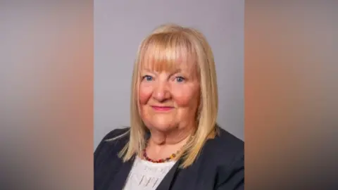 Supplied Liz Maddison has straight blond hair which reaches just below her shoulders. She has fringe and blue eyes. She is wearing a balck jacket, white top and a green, red and yellow necklace.