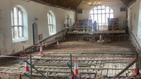 Steve Hubbard/BBC The stage floor, which is long and made of wooden planks. It is covered by other planks and cordoned off by orange cones and tape. White walls with windows flank the stage, and at the end of it people are admiring it from behind a makeshift viewing point.