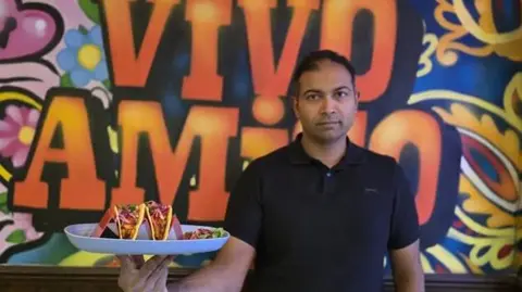 Rajendra Vikram Kupperi Rajendra Vikram Kupperi wearing a black short-sleeved shirt is holding a large white plate with three vibrant tacos. Each taco is filled with colorful ingredients, including shredded lettuce, diced tomatoes, and possibly meat, topped with bright pink pickled onions. The background features a bold, colorful mural with swirling patterns and large orange letters spelling “VIVO AMIGO” against a mix of blue, yellow, and pink designs.