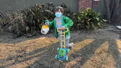 A young boy plays with a toy lawnmower wearing a mask