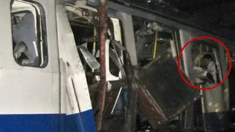 Steven Desborough Steven is highlighted within a computer graphic red circle, stood on a bombed out carriage on the Tube underground. He is not the focus of the picture, but slightly in the background, wearing a shirt and looking down, not in the direction of the camera. Around him is debris, broken doors and loose panels from the explosion on the Tube.