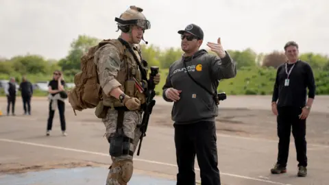 Murray Close/A24 An actor dressed as a contemporary soldier, holding a rifle and wearing a helmet, listens to a film director outside on location