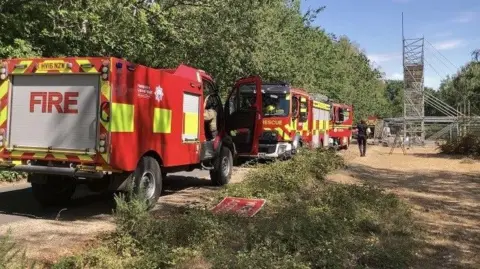 Red fire and rescue vehicles line a tree-bordered lane leading to a scaffold tower and fenced work area in a woodland setting. There is a person in dark uniform in the distance.