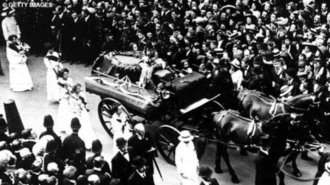 Horses pull the coffin of Emily Davison through a large crowd. There are suffragettes following the procession.