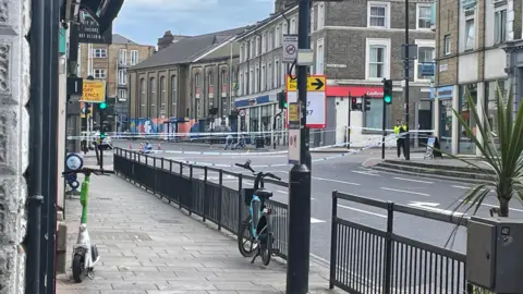 BBC Lavender Road in Clapham at the junction with Wandswoth Road. In the background, forensic officers are seen while police tape is around them. In the foreground is a lamp post, black railings and the pavement and road. 