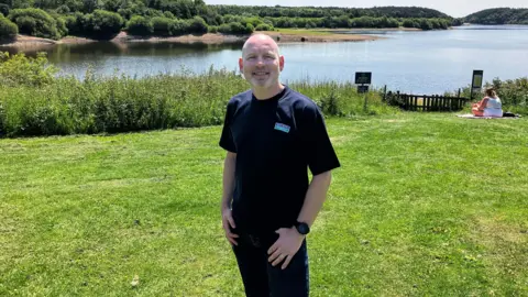 BBC A man wearing a blue T-shirt and jeans stands on grass. There is a reservoir behind him with trees visible on the other side. A woman wearing all white can be seen sitting on the grass behind him to his right.