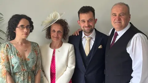 Four members of the Castleton family stand side by side, wearing smart clothes. On the left, a young woman with curly, dark hair and glasses wears a blue floaty dress decorated with orange flowers. Next, an older woman with short, curly brown hair wears a white hat, a bright pink with white polka dots and a white jacket. Beside her, a younger man with short brown hair and short beard wears a navy blue, three piece suit with a gold tie. Beside him, and older man stands, wearing a white shirt, a navy blue waistcoat and a dark purple tie.